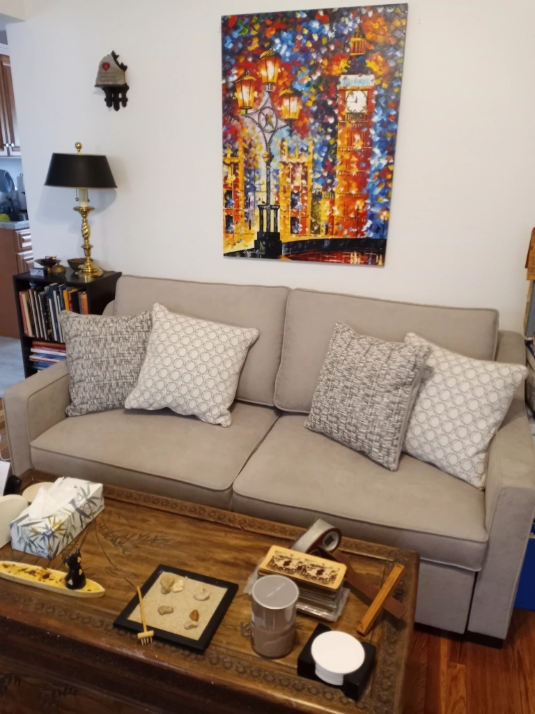 A grey sofa sits against the wall be hind a brown trunk that serves as a coffee table. To its left is a bookcase with a brass lamp on it. Above the sofa is a large semi-abstract painting of Big Ben in riotous colors. 