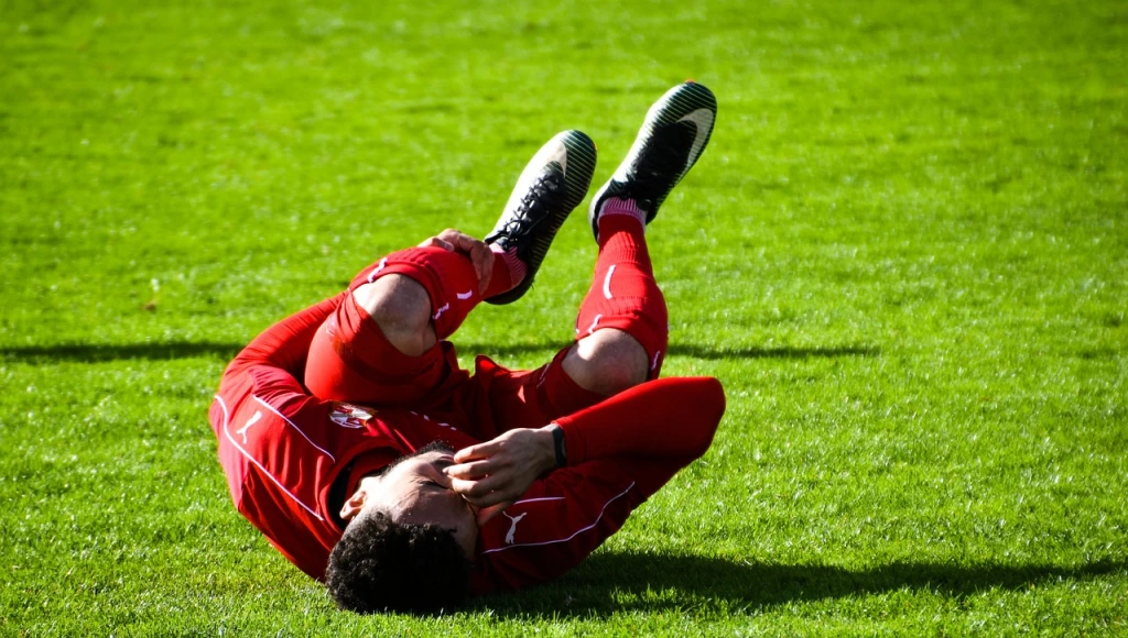 A man dressed in a red soccer uniform writhes in pain while clutching his leg on a green athletic field. 